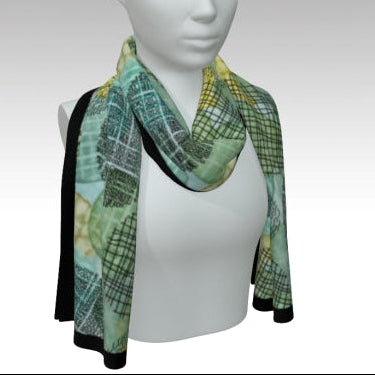 A patterned scarf draped on a white mannequin torso against a gray gradient background. The scarf features a green and black plaid pattern with yellow and green abstract leaf-like shapes and black cuffs.