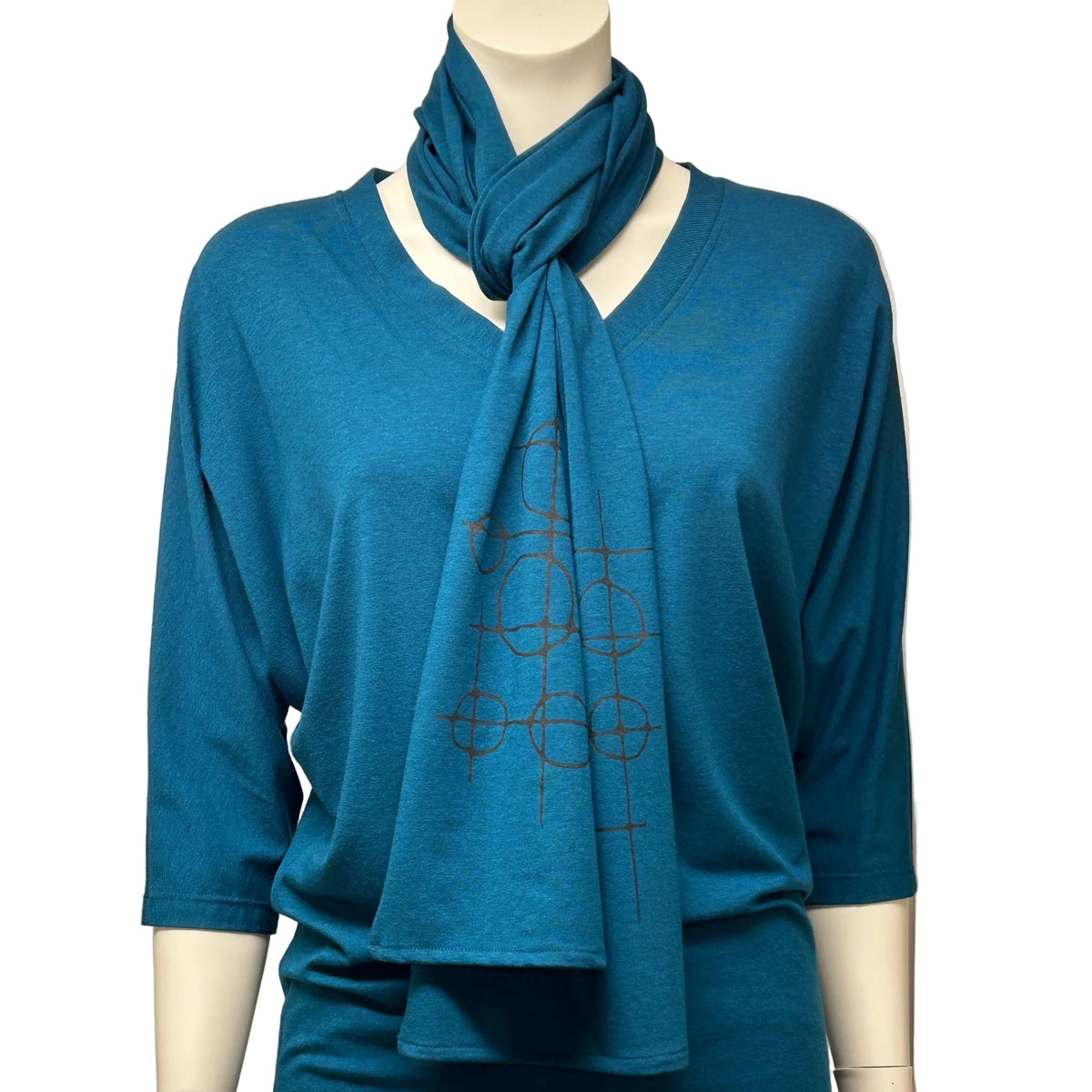 A teal blue knit top and matching scarf displayed on a mannequin, styled with black pants against a neutral indoor backdrop.