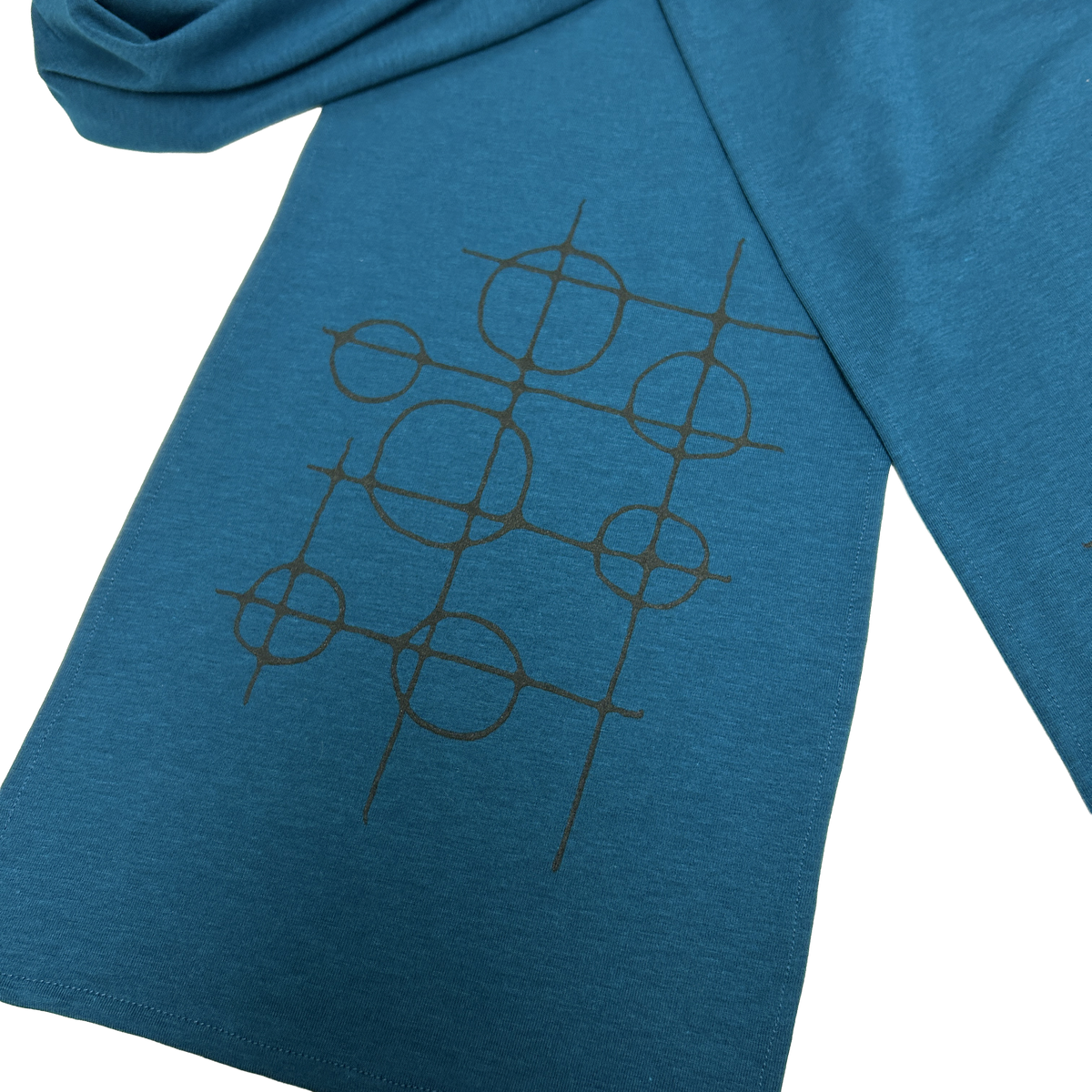 A close-up of a d teal blue garment, scarf, featuring a black geometric line-art graphic of interlocking circles and crosshairs on ends